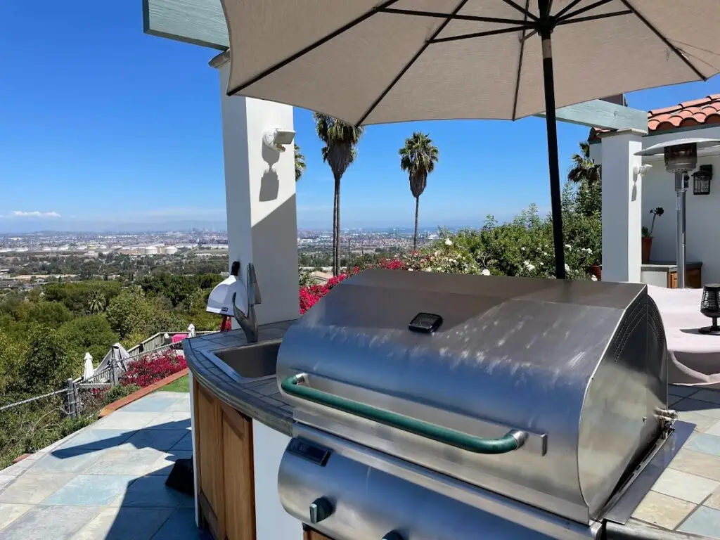 Outdoor patio with a grill under an umbrella, surrounded by lush greenery and vibrant flowers. Overlooks a vast cityscape with clear blue skies.