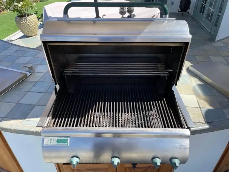 An open stainless steel grill with clean grates on a tiled outdoor patio. Sunlight creates shadows on the surface, and a potted plant is nearby.